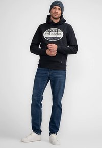 Zwarte hoodie met logo, blauwe jeans en witte sneakers. Stof lijkt zacht. Model draagt ook een blauwe beanie. Simpel ontwerp, casual uitstraling.