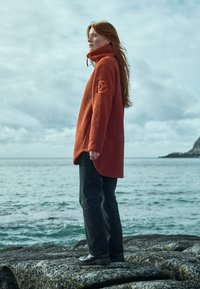 Orange fleece-tröja med hög krage och logotyp på ärmen, tillsammans med mörka byxor och svarta skor, stående på en stenig strand mot vattnet.
