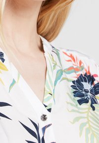 Camisa blanca con un patrón floral en azul, amarillo y verde. Presenta un cuello abotonado y mangas cortas con una textura suave.