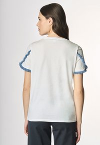 T-shirt bianco con frange blu lungo le maniche. Materiale in cotone morbido, vestibilità regolare e design minimalista. Nessun marchio visibile.