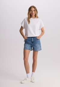 Biała koszulka z krótkim rękawem połączona z jasnoniebieskimi dżinsowymi szortami. Model nosi białe sneakersy i białe skarpetki do kostek. Prosta, codzienna stylizacja.