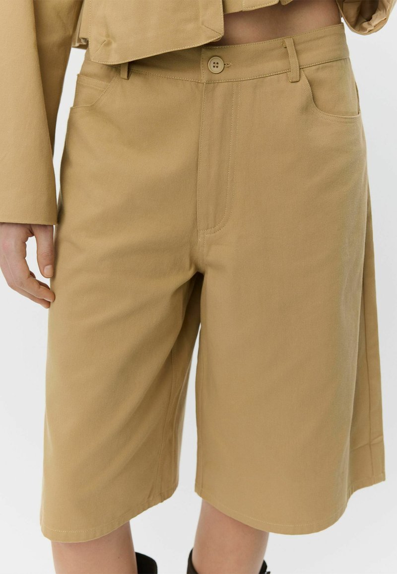 Pantaloncini beige in cotone con vestibilità comoda, lunghezza al ginocchio, dotati di chiusura con bottone, tasche laterali e dettagli cuciti lungo le cuciture.