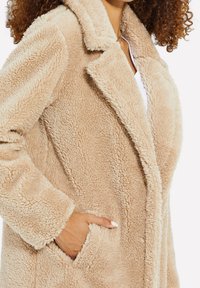 Kvinde iført beige fluffy teddyjakke med hånden i lommen, krøllet hår synligt og hvid top under.
