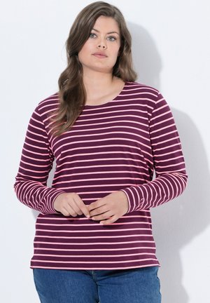 Vrouw met lang bruin haar die een paars en roze gestreept shirt met lange mouwen en een blauwe spijkerbroek draagt, staand tegen een lichte achtergrond.