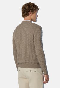 Mann in einem beigen Kabelstrickpullover und hellen Hosen, von hinten gezeigt, vor einem schlichten hellen Hintergrund stehend.