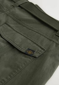 Close-up of olive green fabric with a stitched pocket flap featuring a small black tag with a gold trident logo.