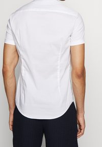 Bílá krátká košile s hladkou texturou, zakulaceným lemováním a jemnými zadními švy, doplněná o tmavé texturované kalhoty.