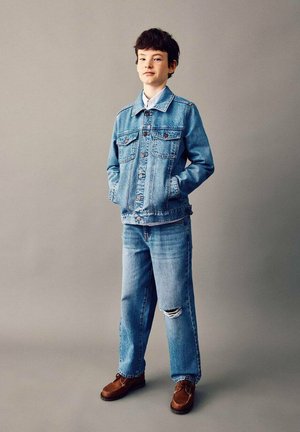 Ragazzo in piedi con le mani nelle tasche che indossa una giacca di jeans azzurro, jeans strappati e scarpe di pelle marrone su uno sfondo grigio uniforme.