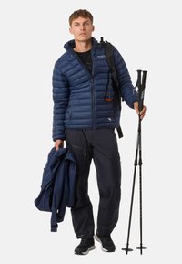 Giacca puffy blu navy con cuciture orizzontali, tasche con zip e un accentuazione arancione. Pantaloni neri e bastoni da trekking, indossati su uno sfondo bianco.