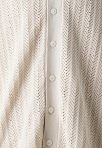 Kremowy kardigan dziany z wzorem jodełki, zapinany na pięć guzików z przodu, o fakturowanym, otwartym splocie.