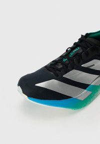 Musta ja hopeanvärinen urheilukenkä mustilla nauhoilla, sinivihreällä pohjalla ja kielessä oleva teksti "ADIZERO" yksinkertaisen vaaleanharmaan taustan edessä.