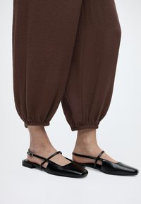 Πόδια με μαύρες μυτερές μπαλαρίνες slingback με λεπτούς ιμάντες, συνδυασμένα με φαρδιά καφέ παντελόνια μαζεμένα στους αστραγάλους.