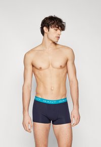Mørkeblå boxershorts laget av myk stoff, med en elastisk midjebånd med turkise "GANT"-logo og en tettsittende design.