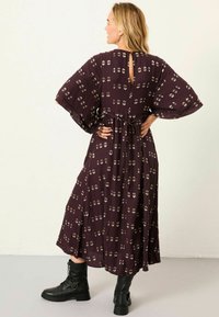 Langes lila Kleid mit floralem Muster, weiten Ärmeln, taillierter Taille und einem Schlüssel-ausschnitt auf dem Rücken, kombiniert mit schwarzen Stiefeletten.