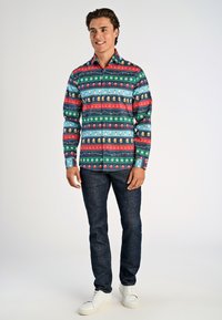Langsleeve shirt in een multicolor feestelijk patroon met groene, rode, blauwe en witte elementen; gecombineerd met donkere jeans en witte sneakers.