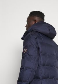 Giubbotto puffer blu navy con cappuccio, caratterizzato da un design trapuntato, una texture liscia e una patch con logo sulla manica.