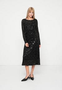 Vestido longo de manga comprida em lantejoulas pretas com um corte ajustado, decote redondo e comprimento até o joelho. Usado com sapatos pretos de bico fino.