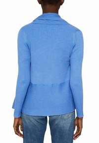 Personne portant un pull côtelé bleu ajusté avec un col montant et un jean bleu, vue de dos sur fond blanc.