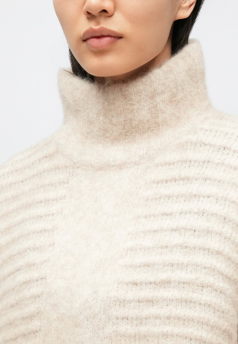 Maglione dolcevita color crema con una morbida maglia testurizzata e dettagli a costine verticali, caratterizzato da un colletto alto e una vestibilità comoda.