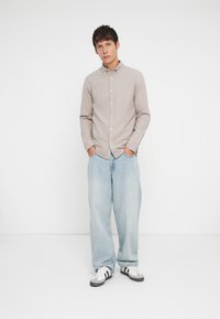 Svetlo béžová košeľa s dlhým rukávom a golierom na gombíky, skombinovaná s voľnými svetlomodrými džínsami a bielymi teniskami s čiernymi akcentmi.