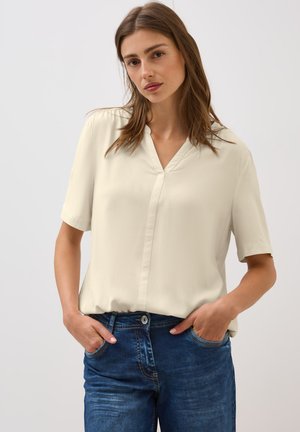Korte mouwen, crème blouse met een V-hals en knoopsluiting, gecombineerd met donkerblauwe jeans met zakken en een lichte vervaging.