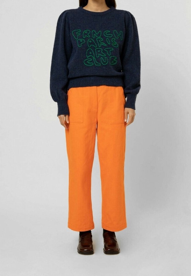 Pull bleu marine avec texte vert « FRENCH PARIS ART CLUB », associé à un pantalon large orange vif et des chaussures marron foncé.