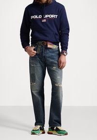 Person som bär en marinblå Polo Sport hoodie, patchwork slitna jeans, en färgglad randig bälte, plaidskjorts ärmar som syns och flerfärgad sneakers.