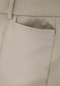 Zbliżenie na kieszeń beżowych spodni z widoczną pętlą na pasek i szczegółowym przeszyciem na gładkim, teksturowanym materiale.