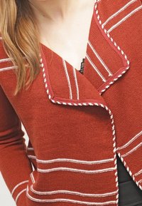 Kardigan barvy rezavé s bílými pruhy, s výstřihem do véčka, pletenou texturou a kontrastním lemováním okrajů.
