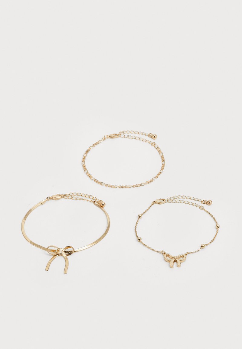 Drie delicate gouden kettingarmbanden, één met een strikcharme, één met een strikvormige band en één met kleine verbonden ovalen.