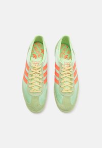 Ett par mintgröna Adidas-sneakers med orangea ränder och beige snören, sedd ovanifrån på en vit bakgrund.