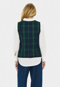 Roheline ja mereväe teksaga vest, millel on vormitud disain. All oleva valge särgi varruka juures on lõikega detail. Mugavad laiad denimpüksid.