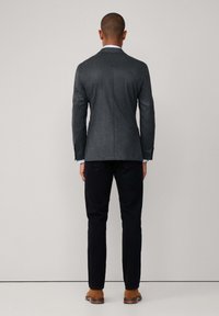 Grau melierter Woll-Blazer mit taillierter Passform, einzelner Schlitze und leicht geschliffenen Revers, kombiniert mit schwarzen Hosen und braunen Wildlederschuhen.