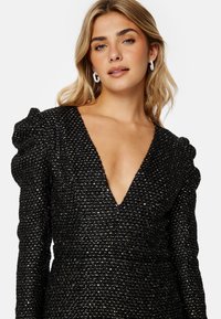 Robe noire texturée avec un décolleté en V profond et des manches bouffantes, mettant en valeur des accents tissés. Modèle accessoirisé avec des boucles d'oreilles blanches en forme de créole.