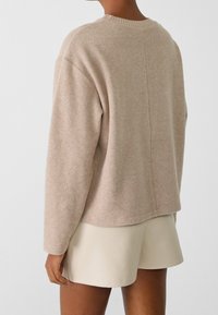 Kvinna som bär en beige långärmad stickad tröja och krämfärgade högmidjade shorts, sedd bakifrån mot en enfärgad bakgrund.