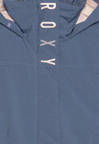 Casaco azul feito de tecido suave, apresentando um logo rosa claro e detalhes em zigue-zague. Inclui capuz e costuras decorativas.