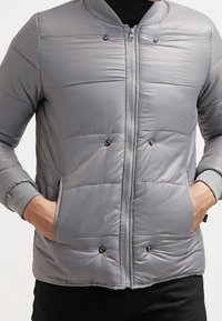 Personne portant une veste rembourrée grise claire avec une fermeture éclair et des poches à boutons-pression, les mains dans les poches, pantalon noir visible.
