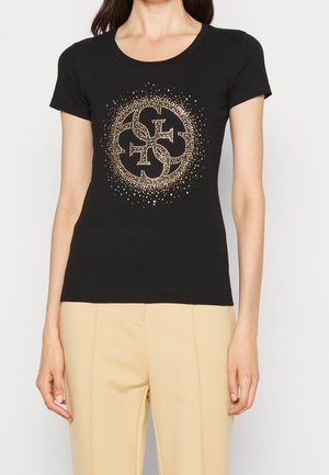 T-shirt noir à manches courtes avec un logo circulaire clouté doré au centre, en coton doux. Associé à un pantalon beige.