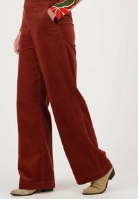 Wijdvallende corduroy broek in roestkleur met verticale ribbels. Voorzien van zakken en een gladde textuur, gecombineerd met lichtgekleurde enkelboots.