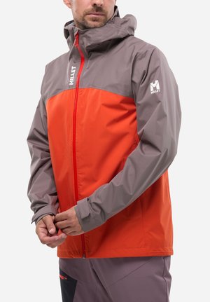 Homme portant une veste d'extérieur Millet bicolore gris et orange avec capuche, ajustant le poignet sur un fond clair.