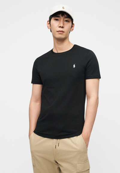 Μαύρο βαμβακερό T-shirt με κοντά μανίκια και στρογγυλή λαιμόκοψη, με μικρό λευκό λογότυπο στο στήθος. Συνδυάζεται με χακί cargo σορτς και μπεζ καπέλο.