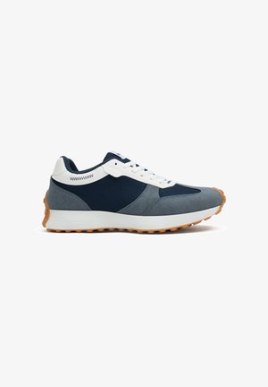Zapatilla baja con malla azul marino, superposiciones de ante gris, cordones blancos y suela de goma color chicle, mostrada desde el lado exterior sobre fondo blanco.