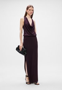 Μακρύ, σκούρο μωβ φόρεμα με κρεμαστή λαιμόκοψη και πλαϊνό άνοιγμα, σε συνδυασμό με μαύρη τσάντα και ψηλές γόβες, από λεία υφάσματα.