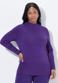 Žena oblečená v fialovém svetr s dlouhým rukávem, zdobeném diamantovými vzory a malými kamínky, v kombinaci s odpovídajícími fialovými kalhotami, stojící proti bílé zdi.