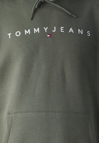 Zelená mikina s kapucňou, predným vreckom, sťahovacou šnúrkou a vyšívaným logom "TOMMY JEANS" s červenými a bielymi akcentmi na hrudi.