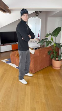 Μαύρο ριγωτό πουλόβερ με φερμουάρ, γκρι παντελόνι σε φαρδιά γραμμή, λευκά αθλητικά παπούτσια, καπέλο με σχέδια; Εσωτερικός χώρος με ξύλινο δάπεδο και γλάστρα.