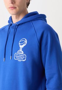 Sudadera de algodón azul con un gráfico frontal que presenta un trofeo y un balón de baloncesto. El texto incluye "Tour del Campeonato de Lower Manhattan 11 de marzo de 1990."
