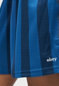 Μπλε και ναυτικό κάθετα ριγωτά σορτς από διχτυωτό ύφασμα με μικρό λογότυπο "obey" κοντά στο τελείωμα, που φοριούνται από ένα άτομο.