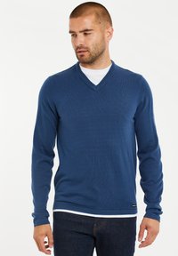Blauer V-Ausschnitt-Pullover aus weichem Strickmaterial, mit langen Ärmeln und einem dezenten Logo-Akzent am Saum. Über einem weißen Hemd getragen.