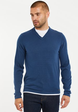 Blauwe V-hals trui gemaakt van zachte breimateriaal, met lange mouwen en een subtiele logo-accent aan de zoom. Draagt over een wit shirt.